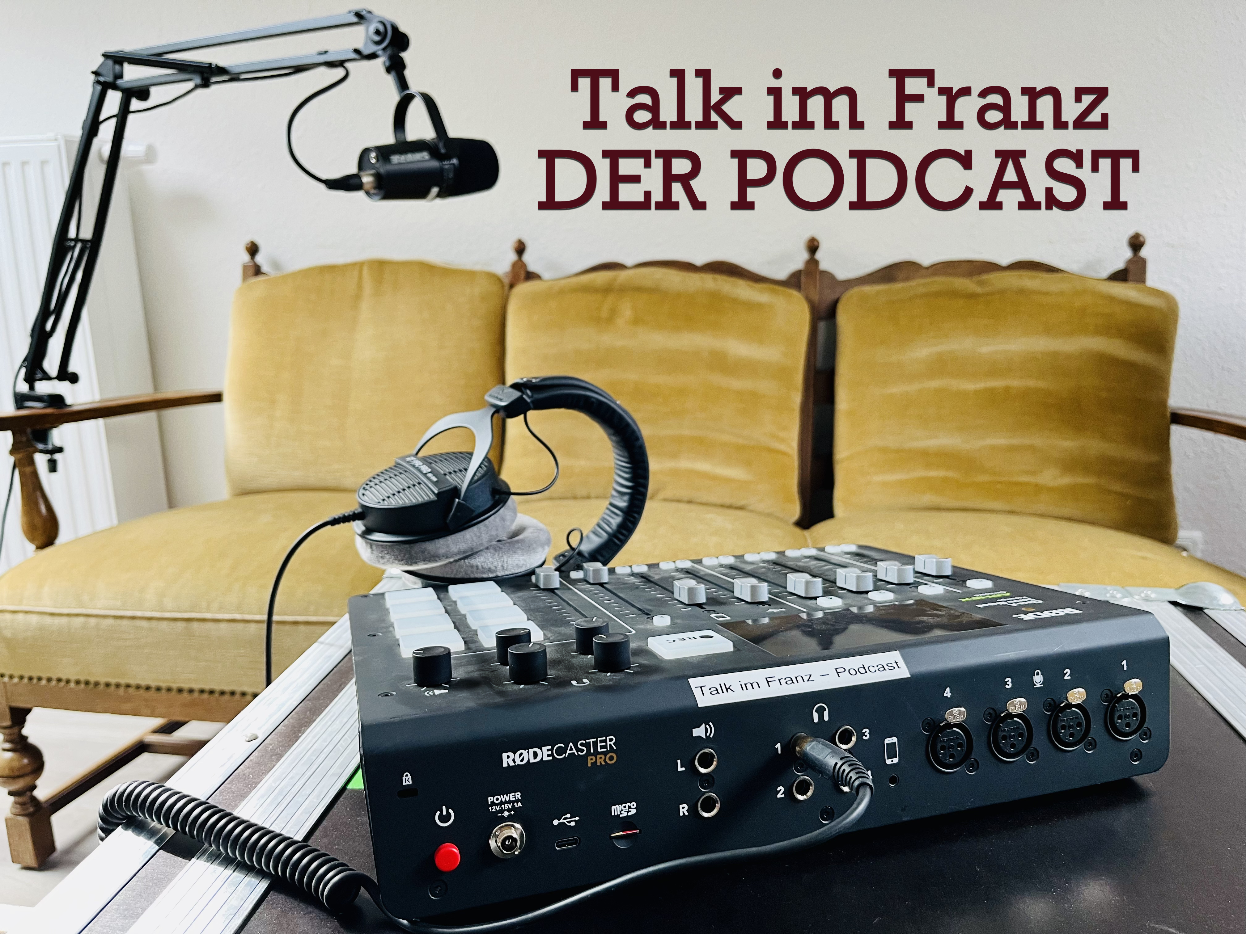 Das Bild zeigt ein Podcast-Aufnahme-Setup. Im Vordergrund befindet sich ein RODECaster Pro Audio-Mixer mit zahlreichen Knöpfen, Tasten und Anschlüssen. Auf dem Mixer liegt ein Kopfhörer. Links im Bild ist ein Mikrofon an einem verstellbaren Mikrofonarm befestigt. Im Hintergrund steht ein gelbes, gepolstertes Sofa mit hölzernen Armlehnen und einer dekorativen Rückenlehne. An der Wand über dem Sofa ist der Schriftzug „Talk im Franz – DER PODCAST“ zu sehen.